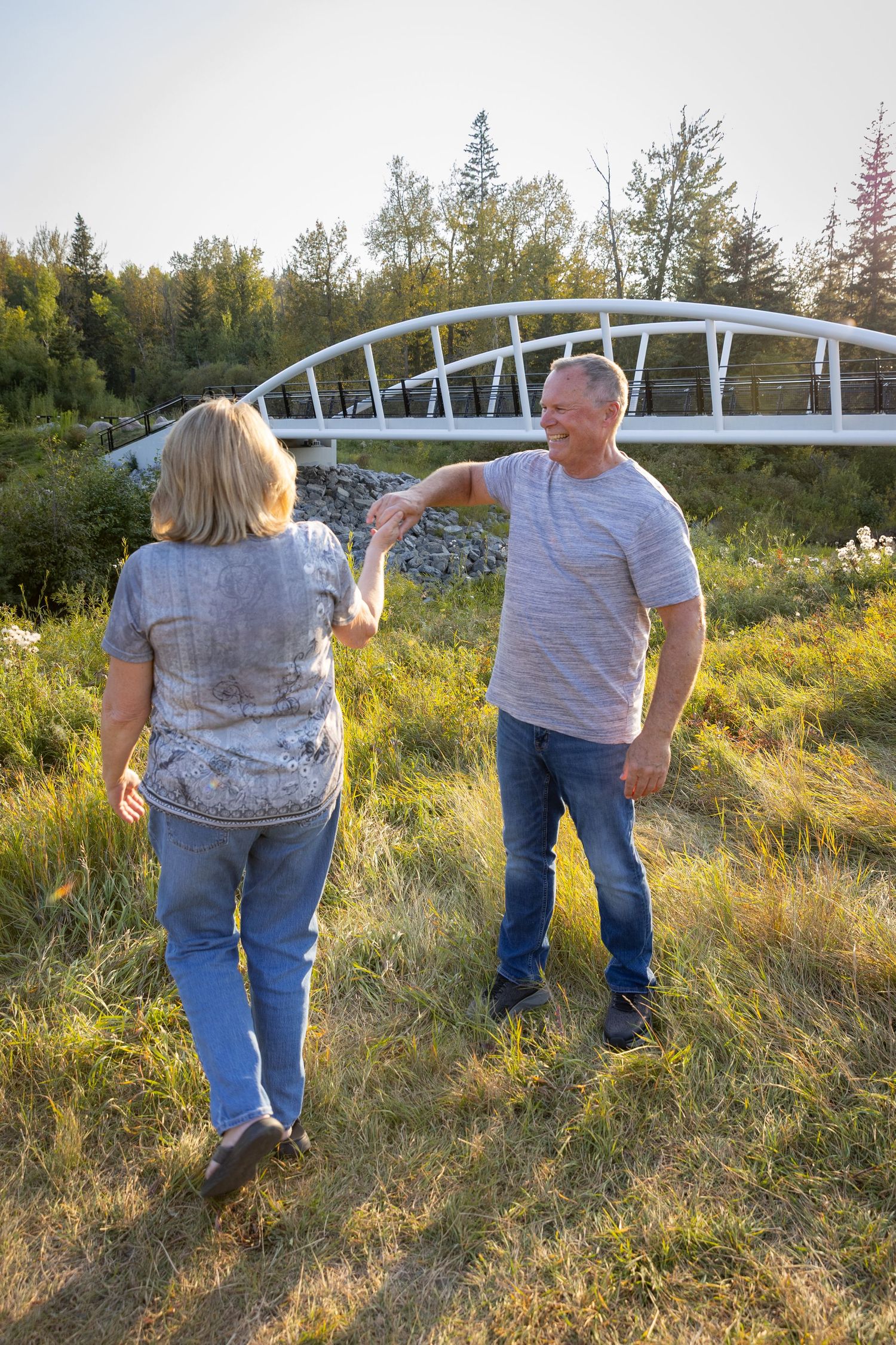 Mature couple holding hands and dancing in a sunny green field near a white bridge.