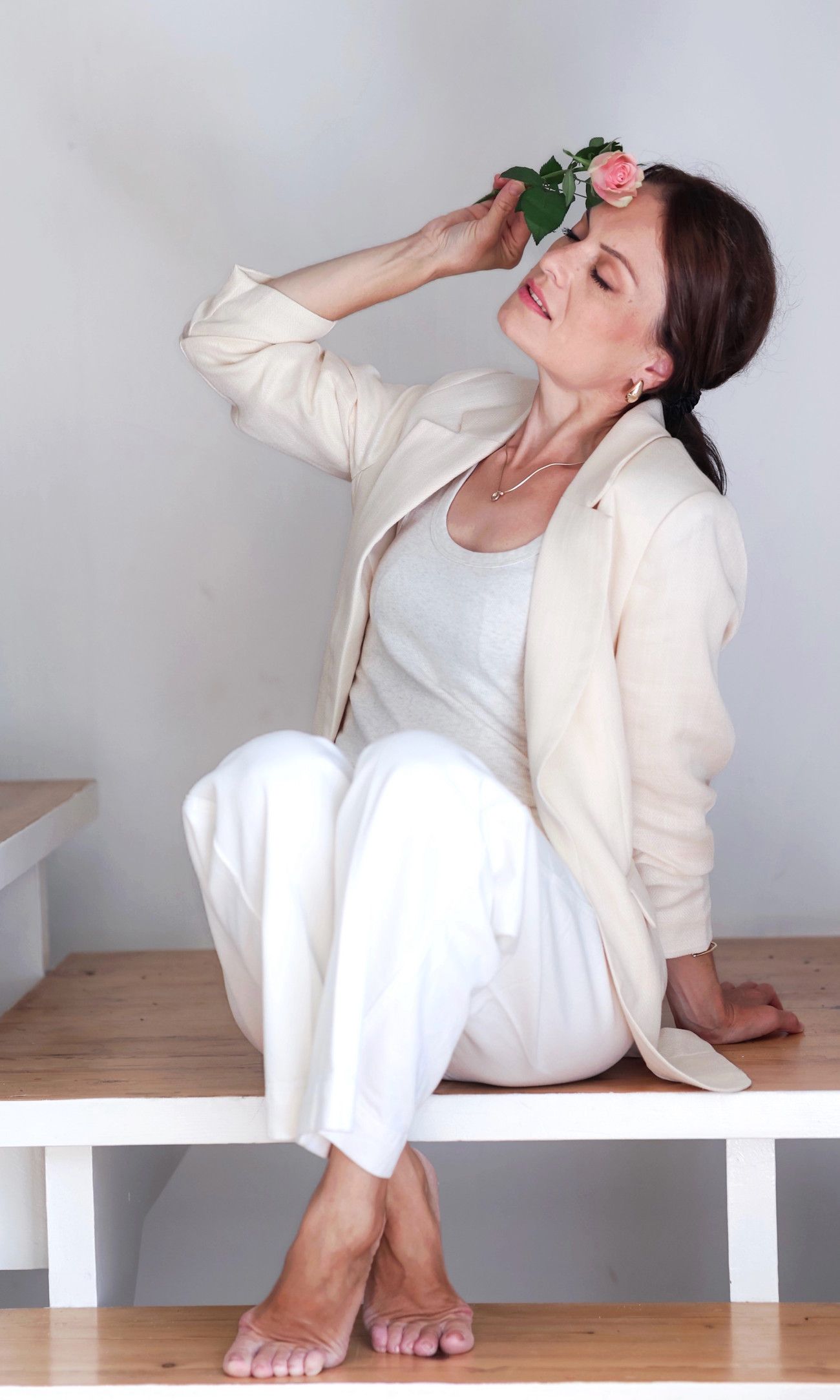 Barefoot woman in cream blazer and white pants sits on bench holding a pink rose to her head.