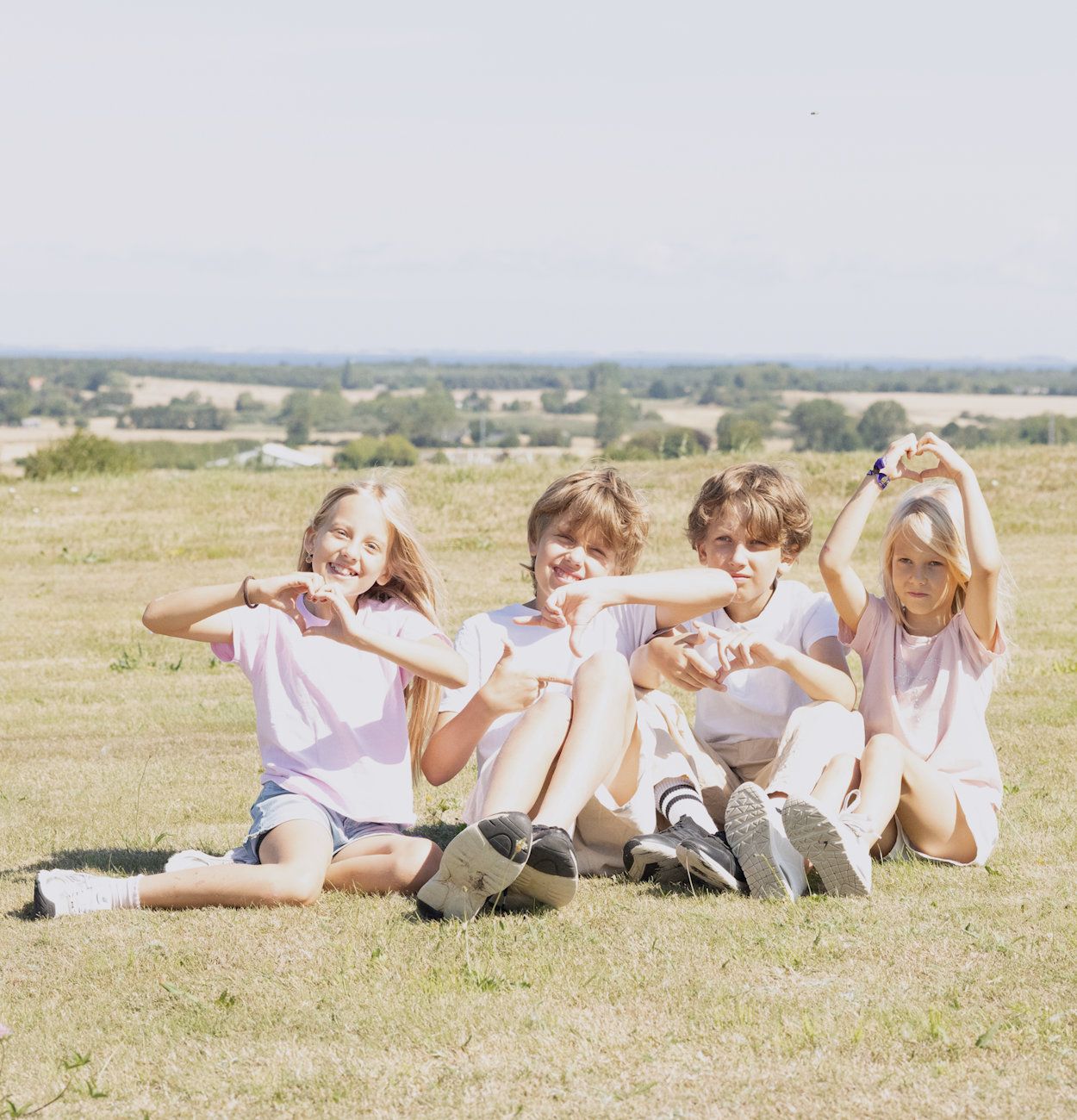 Four children sitting on grass in a sunny field, making heart shapes with their hands and smiling.