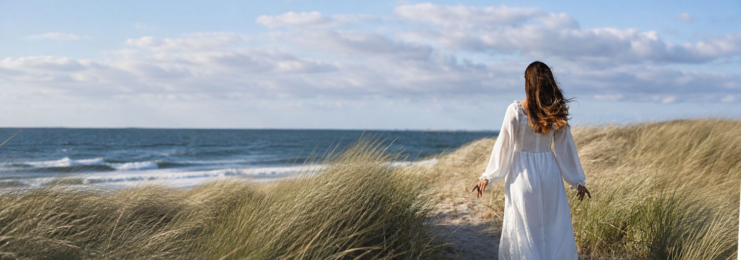 Woman in white dress walking through beach dunes with ocean waves and cloudy sky in the background.