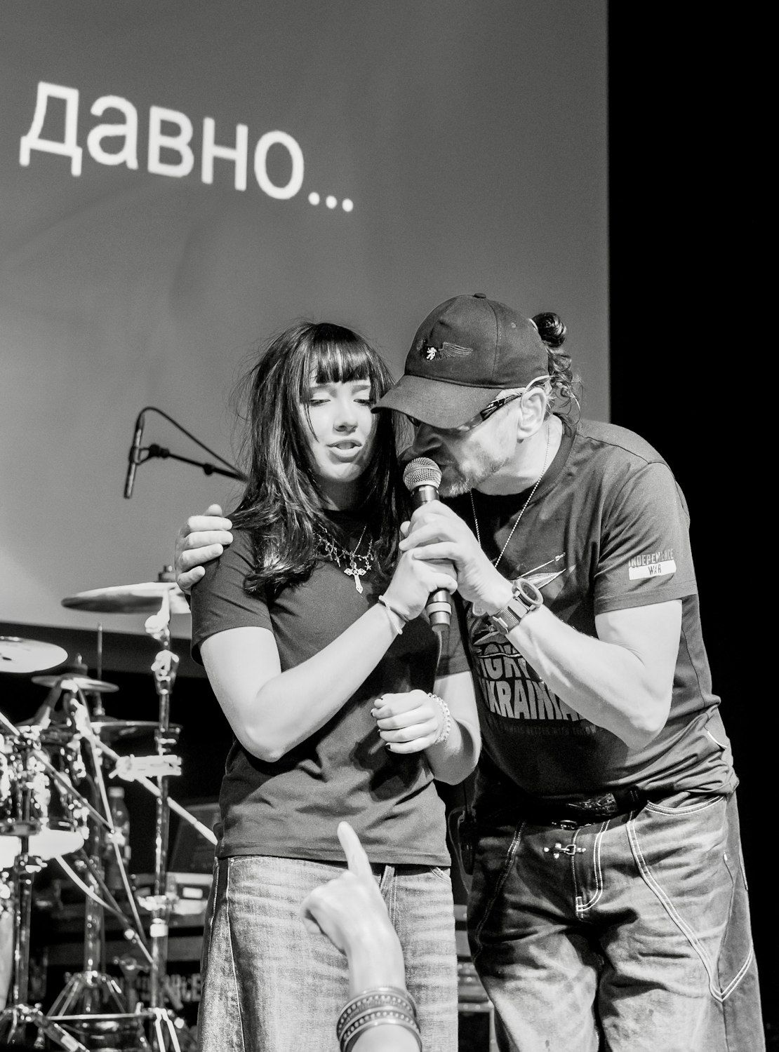 Black and white photo of two performers on stage, man in cap sharing microphone with woman wearing cross necklace.