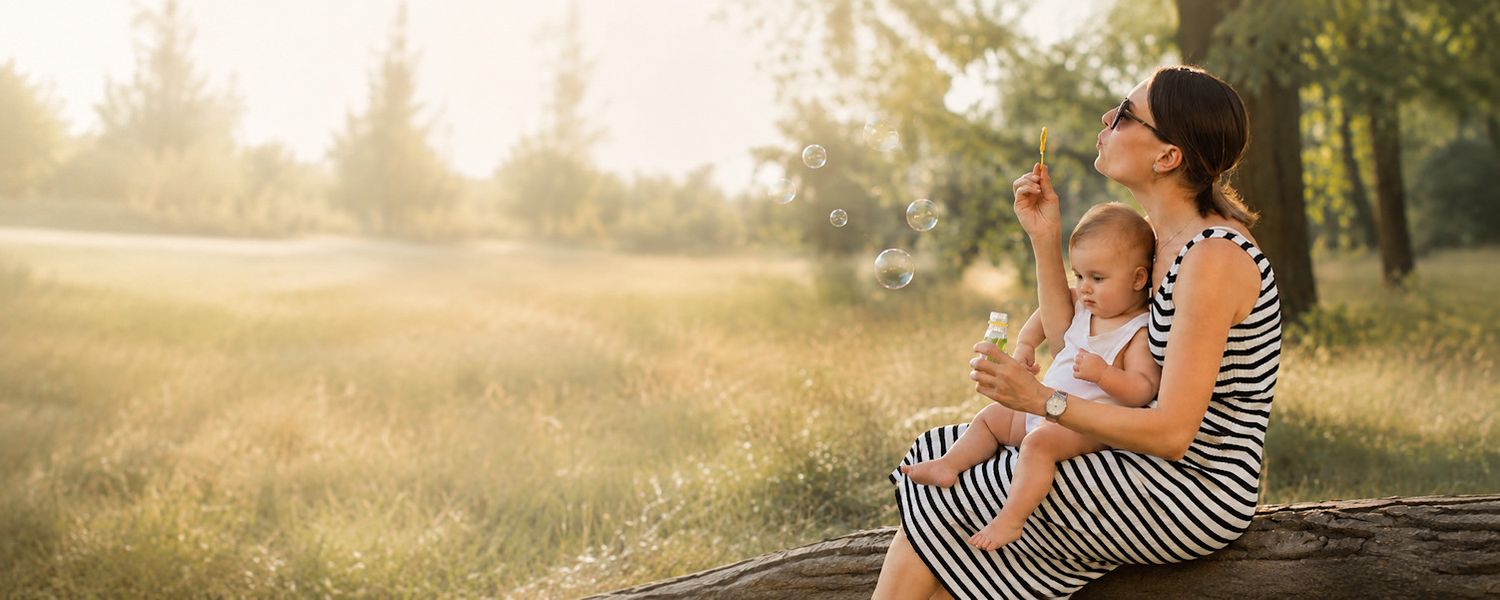 Mother and baby sitting on a log in a sunny park, blowing bubbles together in matching striped dresses.