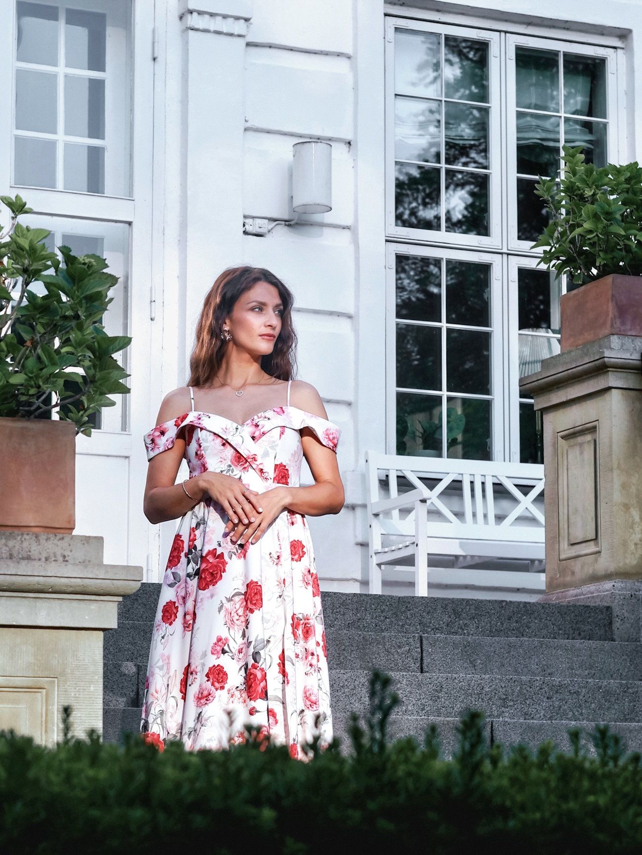Woman in floral off-shoulder red and white dress stands on steps outside elegant white building.