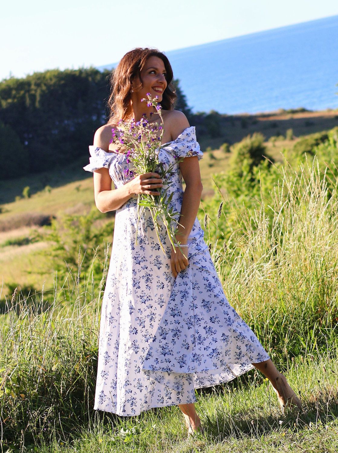 Woman in floral maxi dress holding wildflowers in green coastal meadow.
