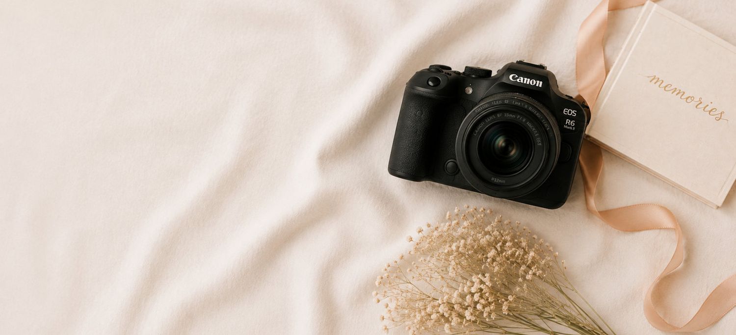 Canon DSLR camera with 50mm lens on white fabric beside dried flowers and pink ribbon.