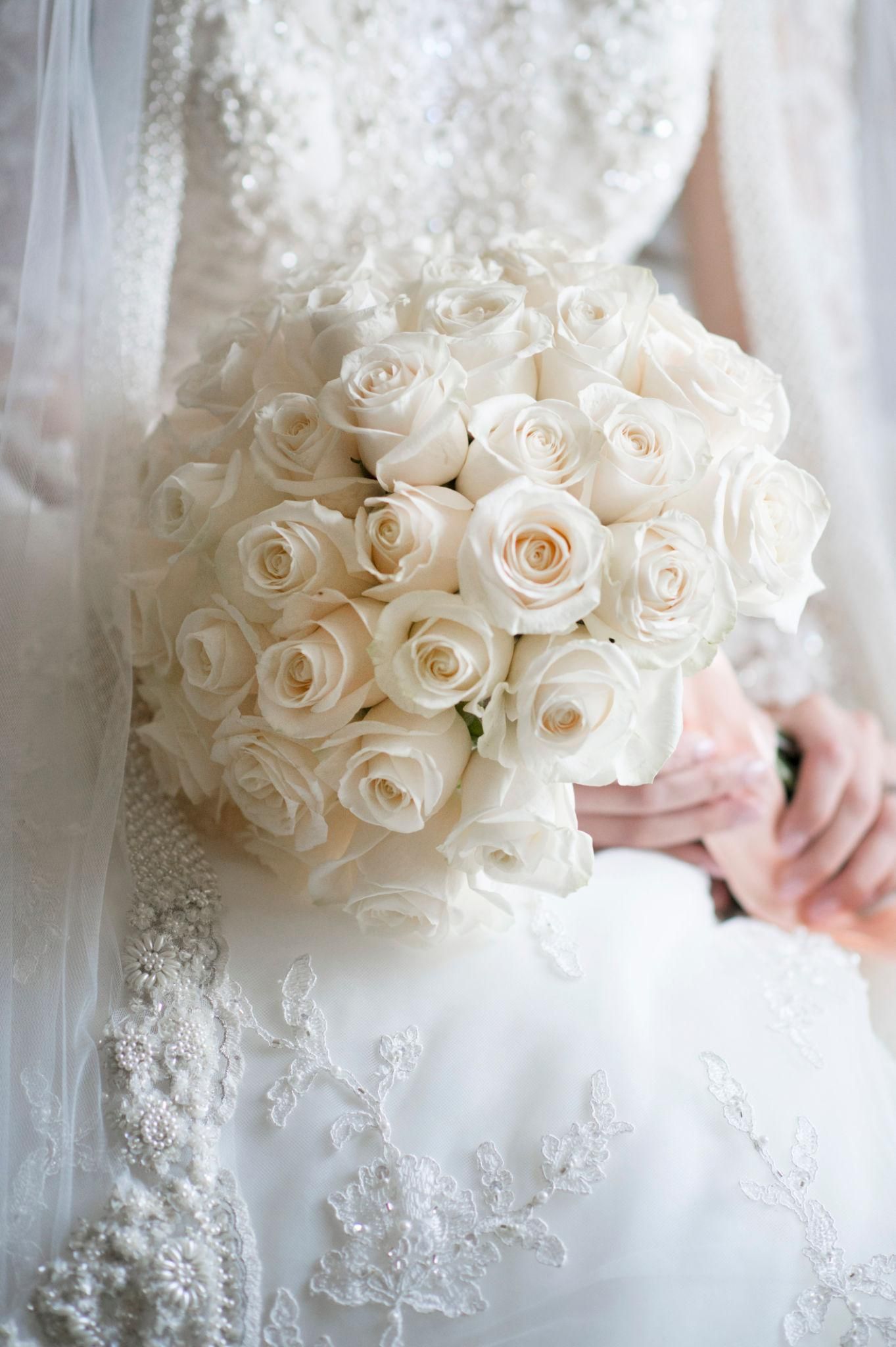 A bride in a lace gown holds a lush round bouquet of cream and white roses against her wedding dress.