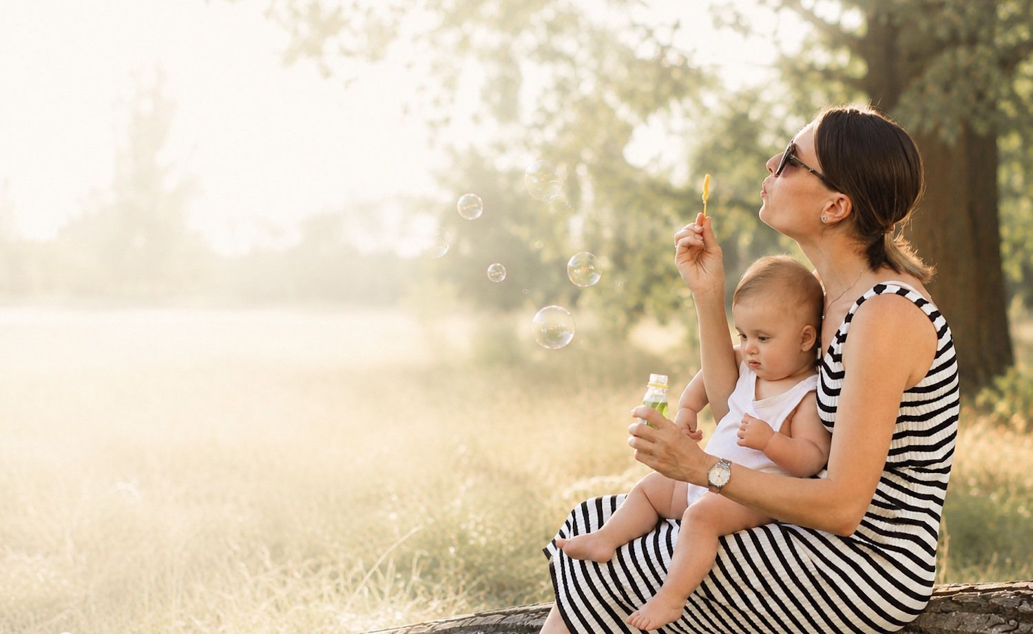 Mother in striped dress blows dandelion with baby on lap in sunny meadow setting.