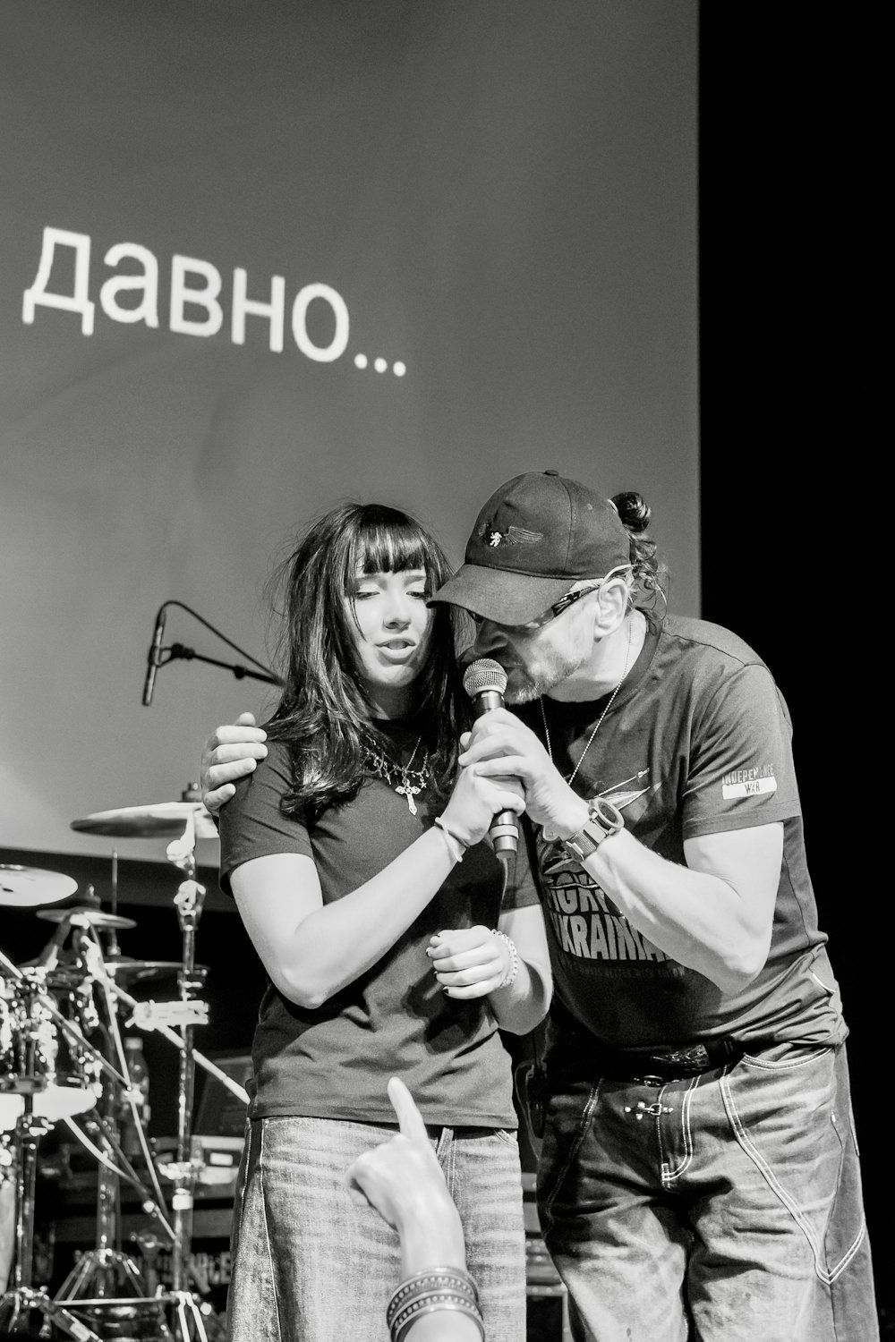 Two performers share a microphone on stage in black and white, with Cyrillic text visible in the background.