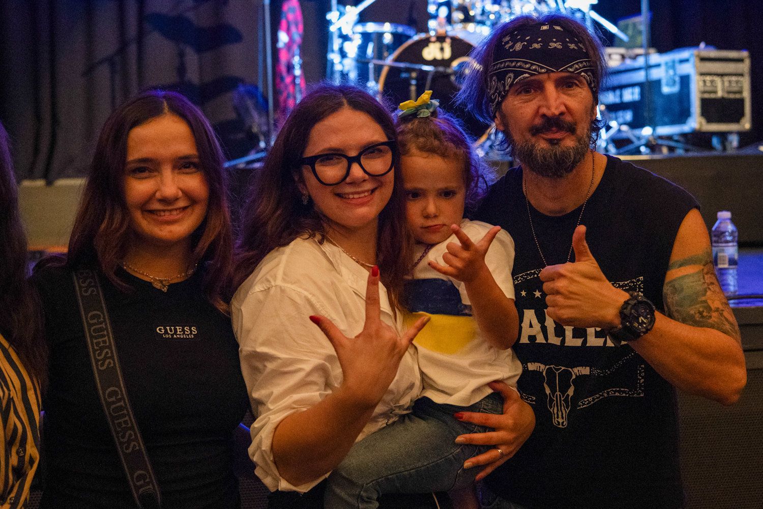 Family of four posing with rock hand gestures at a live music concert venue with stage lights.