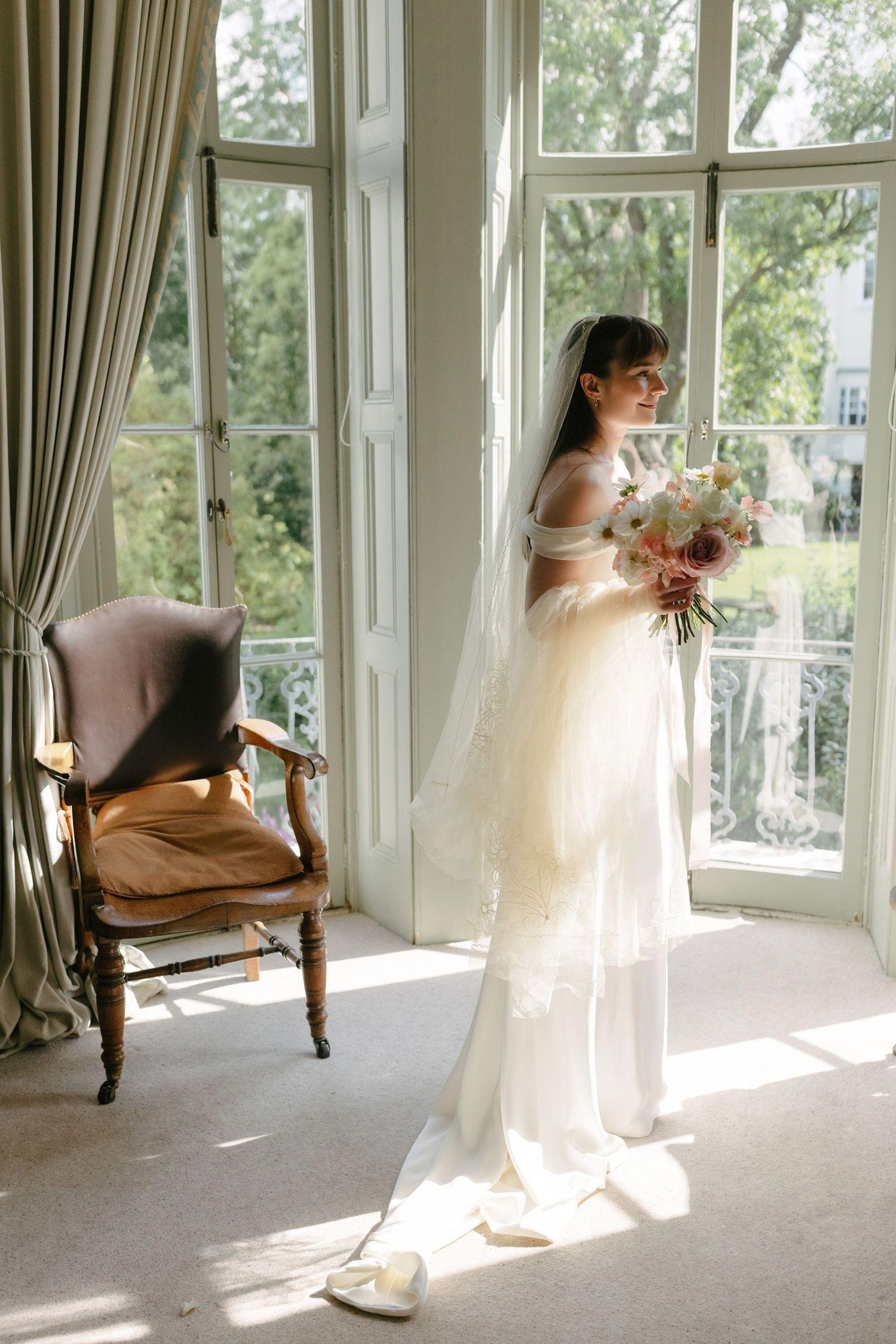 Elegant bride in a white gown and veil stands by a sunlit French window holding a bouquet.
