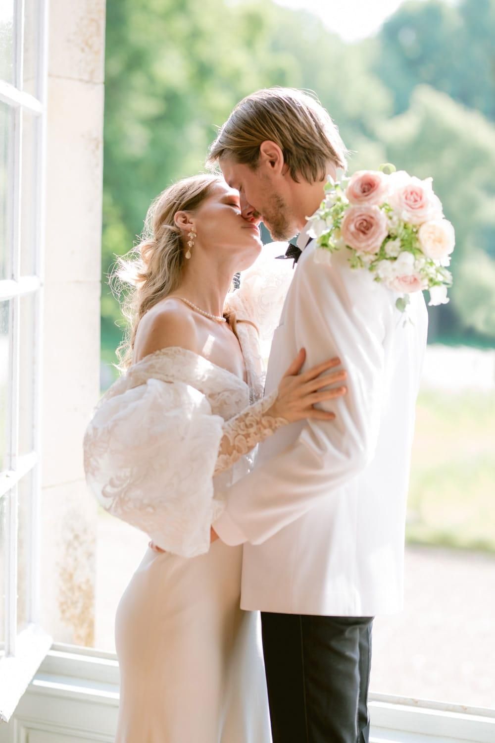 Couple shares a kiss by a bright window, bride in lace gown holding pink roses bouquet.
