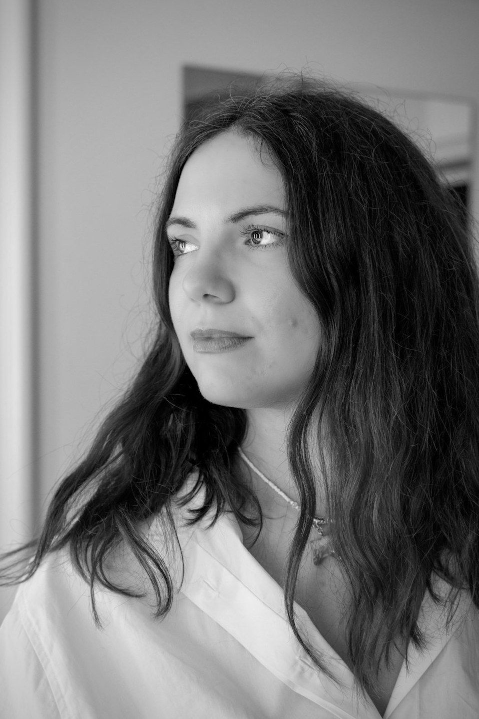 Black and white portrait of a young woman with long dark hair wearing a white shirt, gazing thoughtfully to the side.