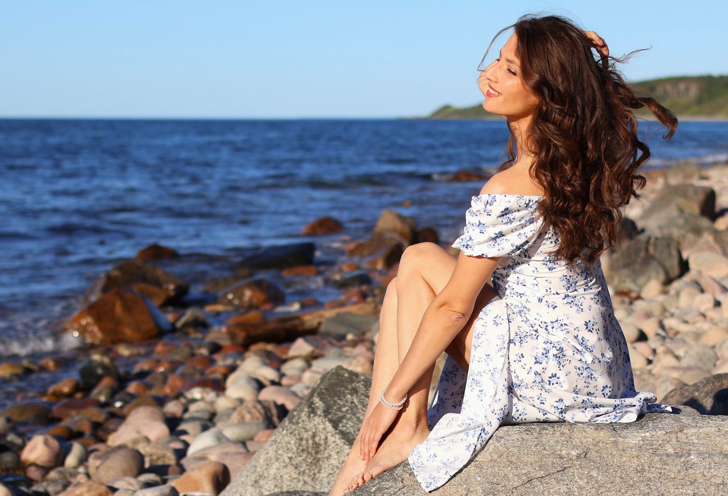 Woman in floral dress sitting on rocky beach enjoying sunny day.