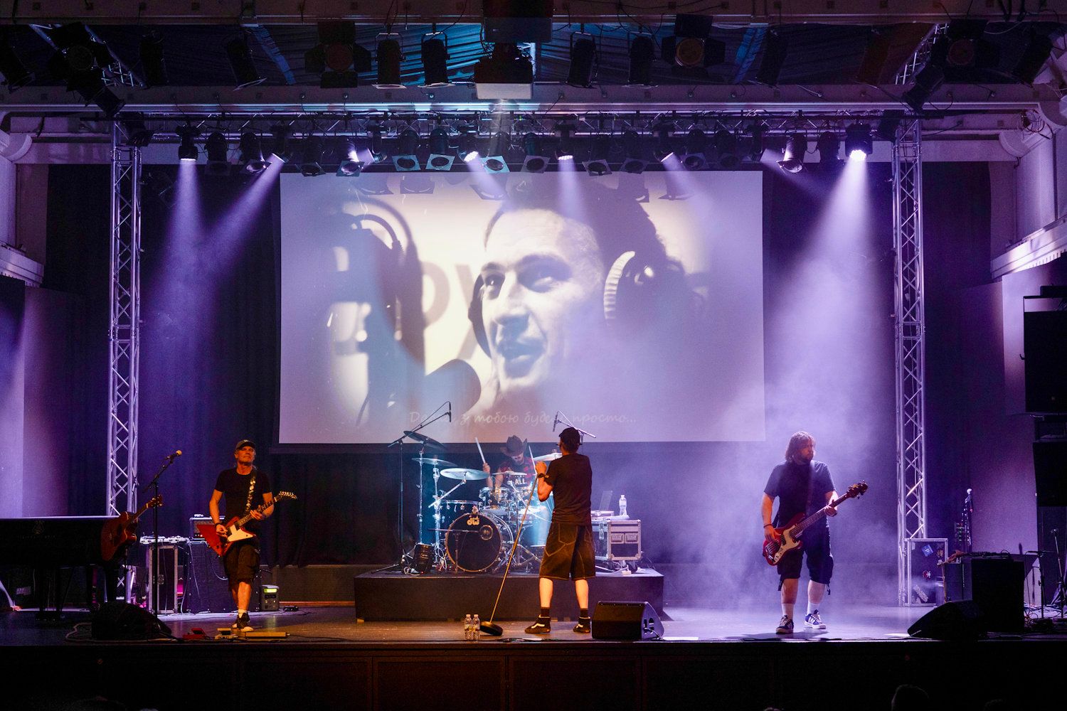Live band performs on stage with dramatic lighting and large projection screen displaying a close-up face behind them.