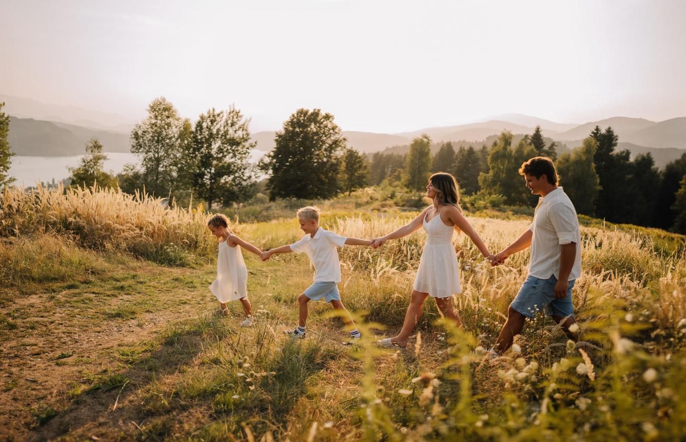 Happy family holding hands and walking through golden meadow with mountain lake view.
