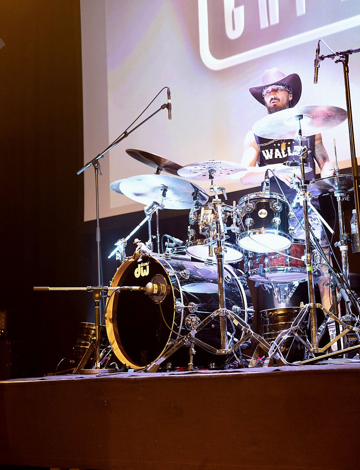 Drummer performing on stage with DW drum kit, wearing cowboy hat and tank top under concert lighting.