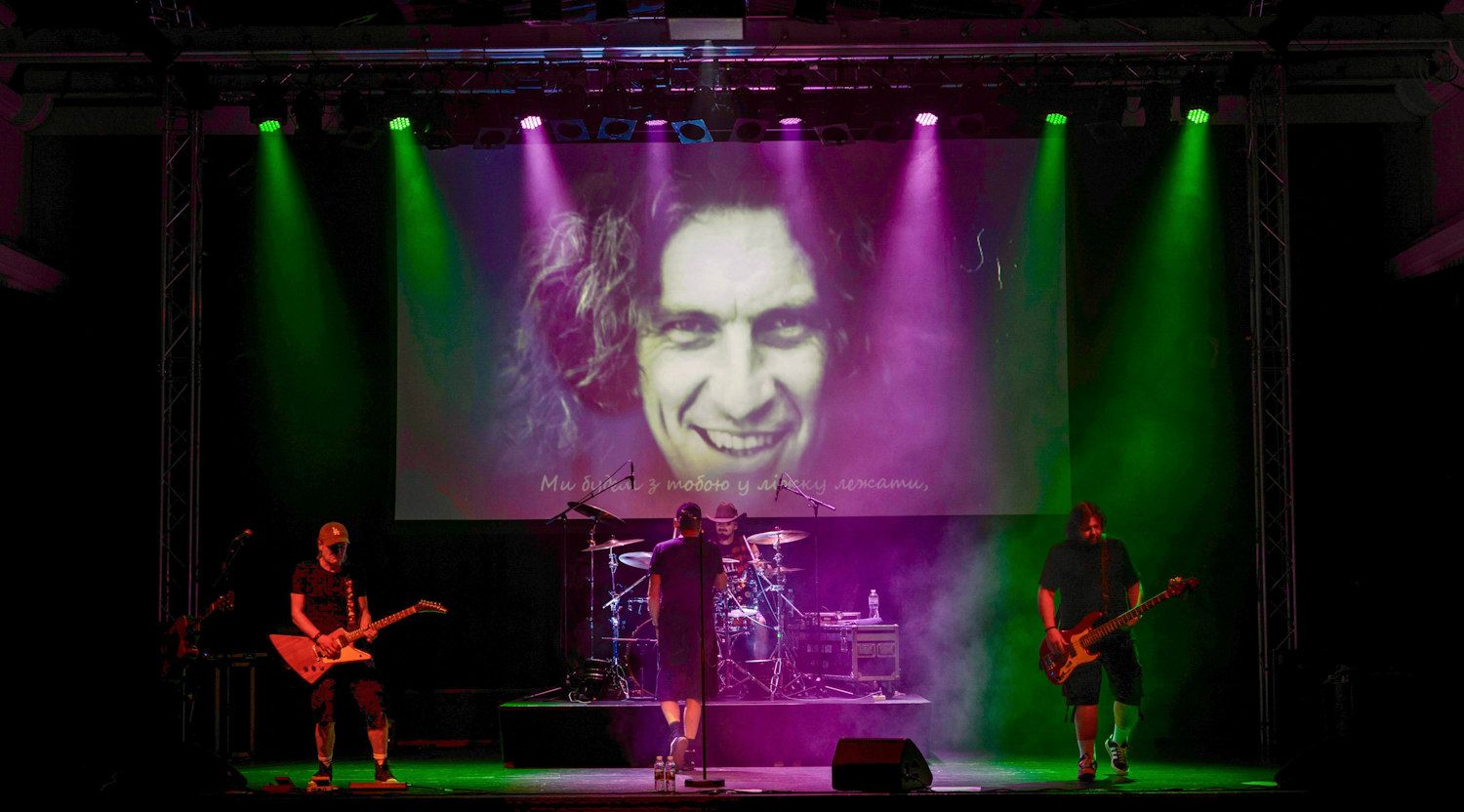 Rock band performs on stage with dramatic green and pink lighting, large screen displaying a man's face behind them.