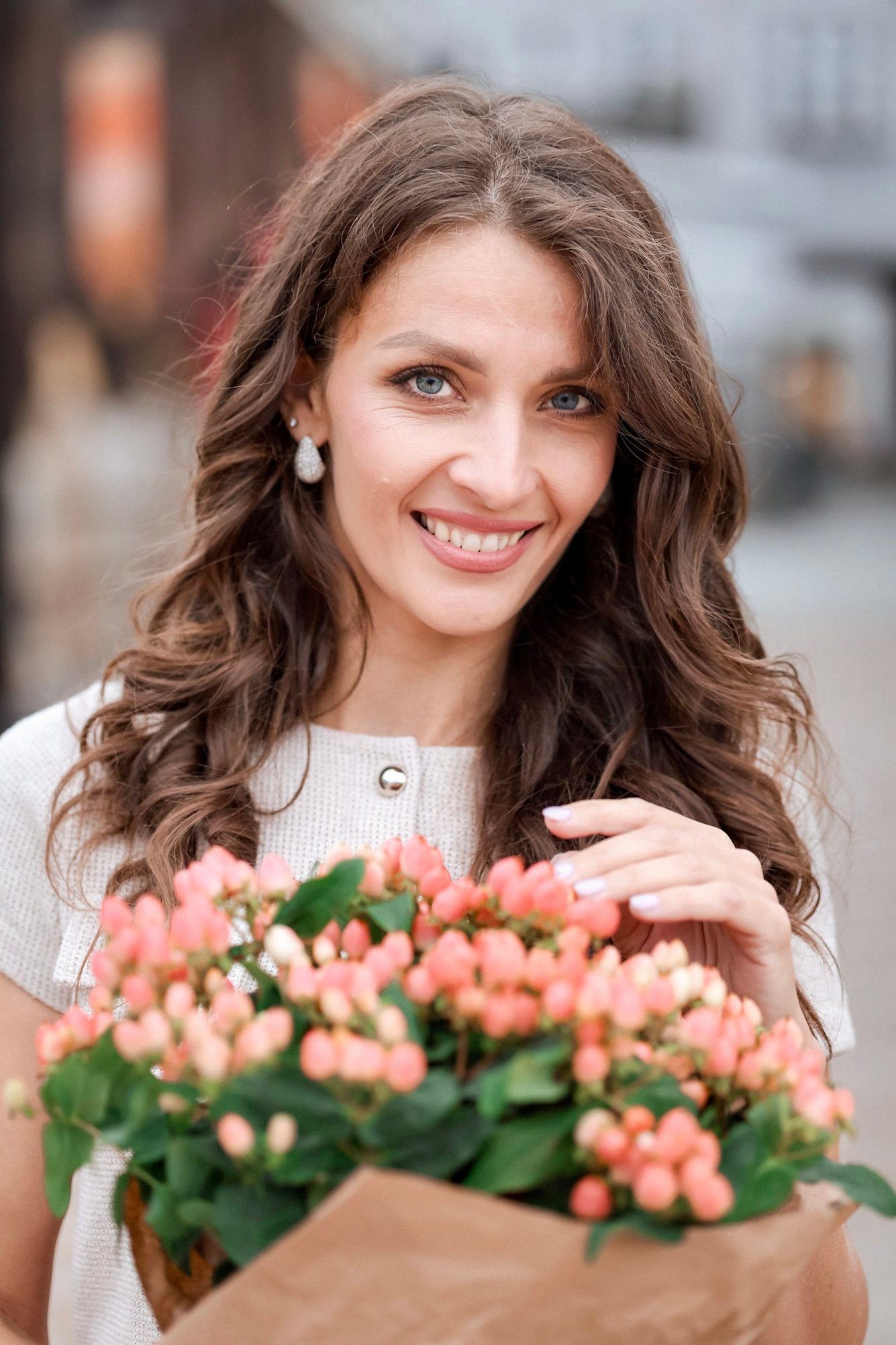 Smiling brunette woman holds a bouquet of pink flowers while standing outdoors on a city street.