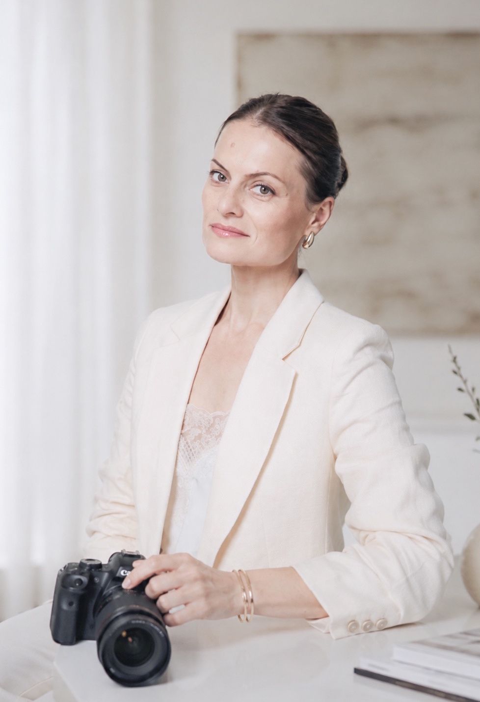 Professional woman in cream blazer holds DSLR camera while seated at a bright white desk.