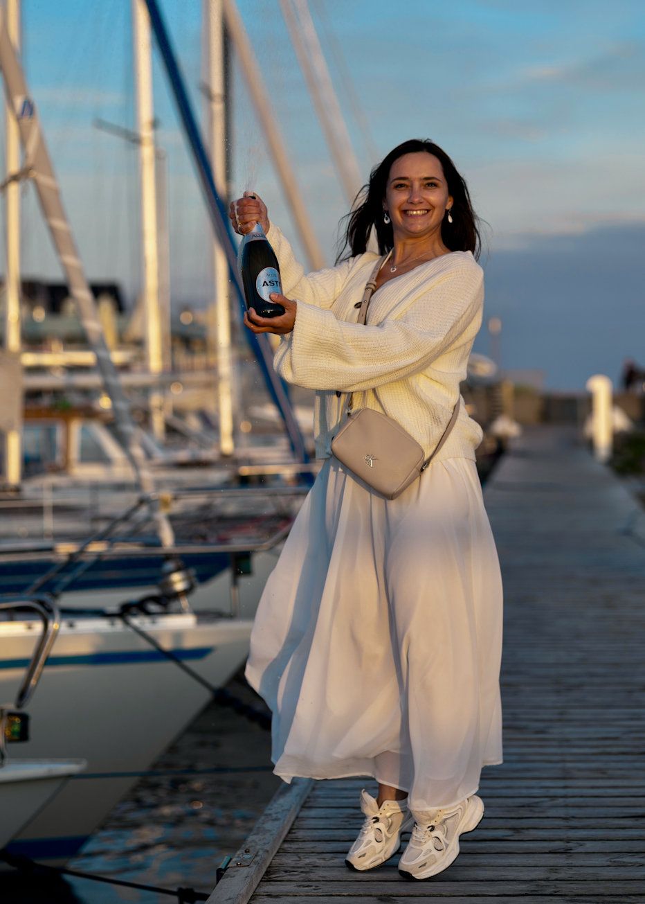 Woman in white dress holding champagne bottle at marina dock with sailboats at sunset.