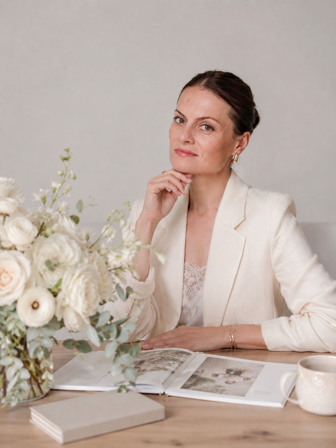 Elegant woman in cream blazer sits at desk with white floral arrangement and open photo album.