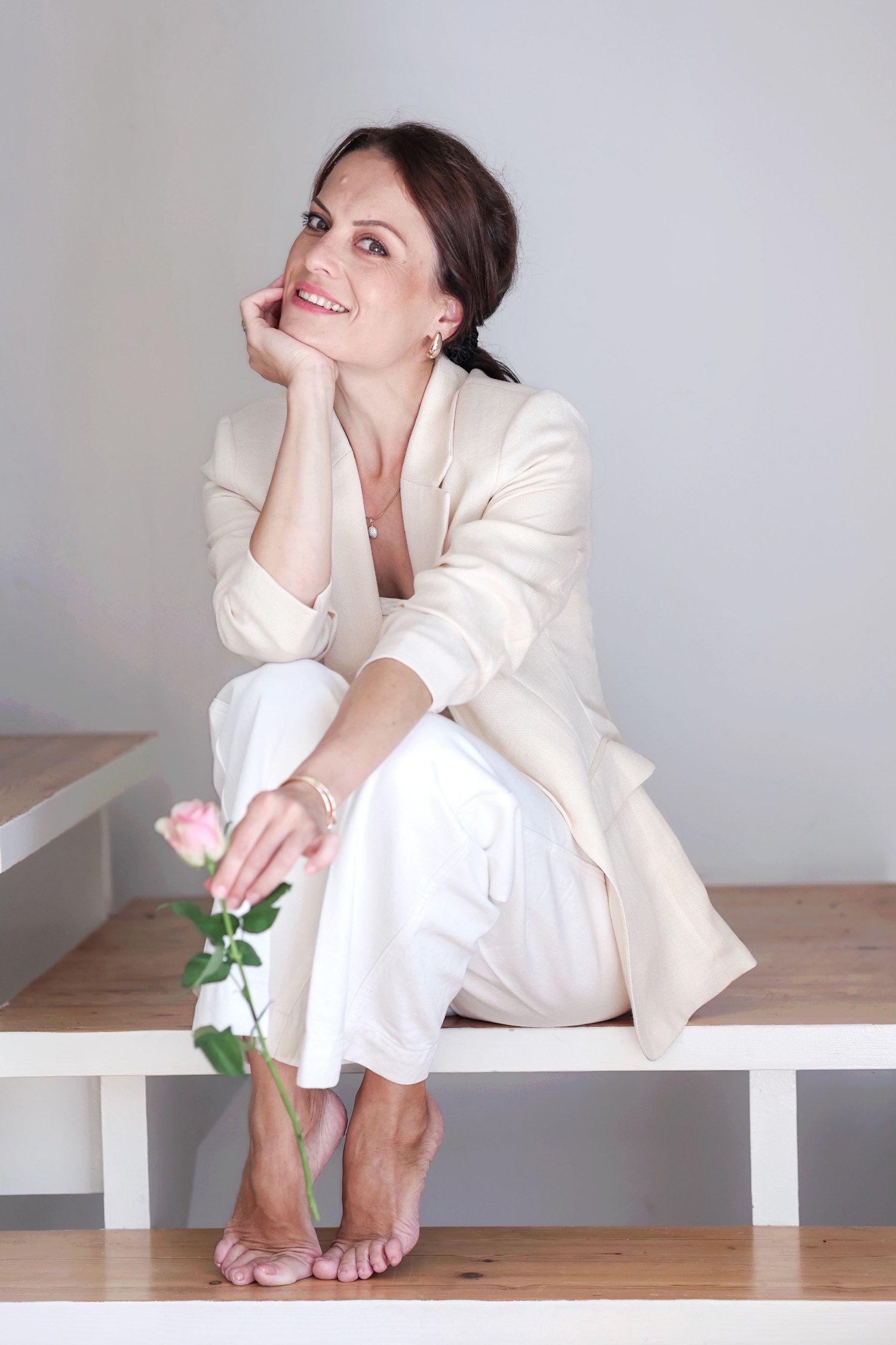 Smiling woman in white blazer and pants sits barefoot on bench holding a pink rose.