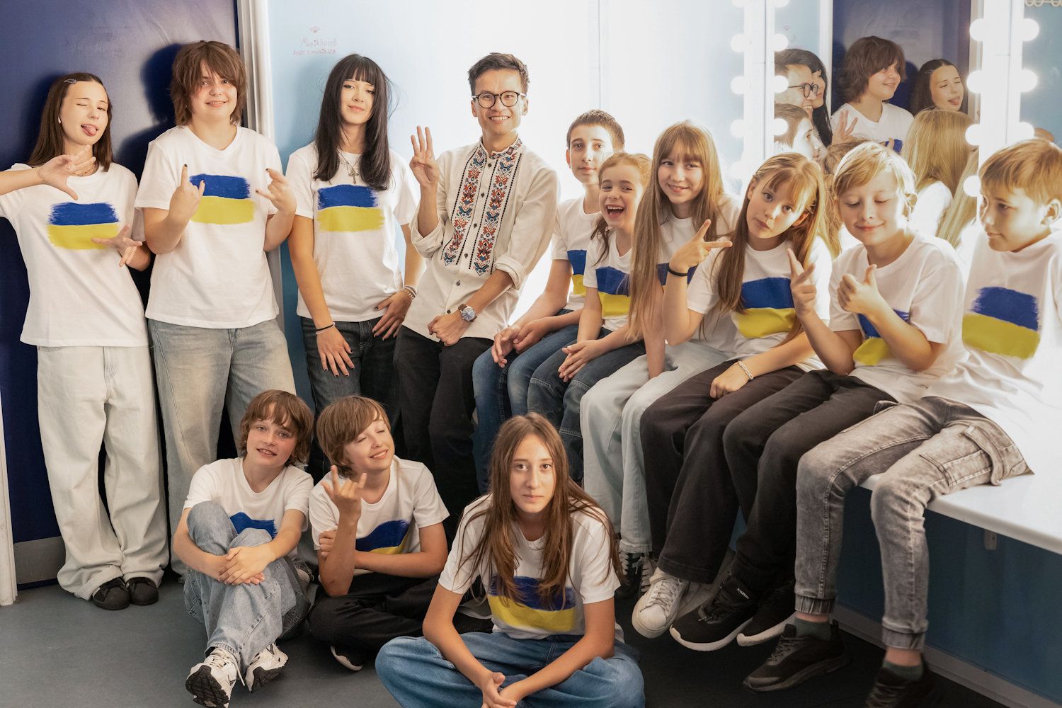 Group of teens in white shirts with Ukraine flag patches posing together in a bright dressing room.