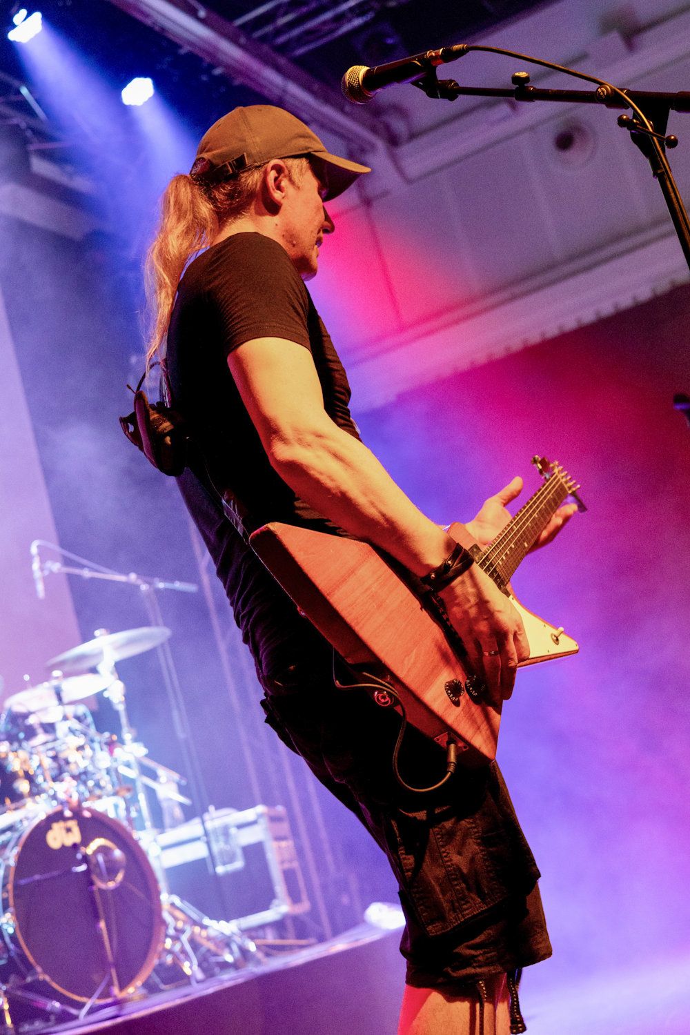 Male guitarist with long hair performs on stage under purple and blue lights with drum kit visible in background.