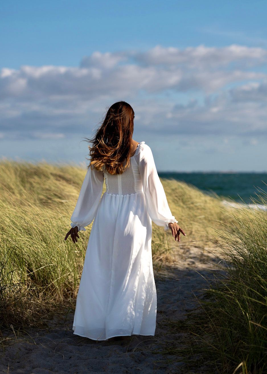 Woman in white maxi dress walking on sandy beach path through grass dunes toward ocean under blue sky.