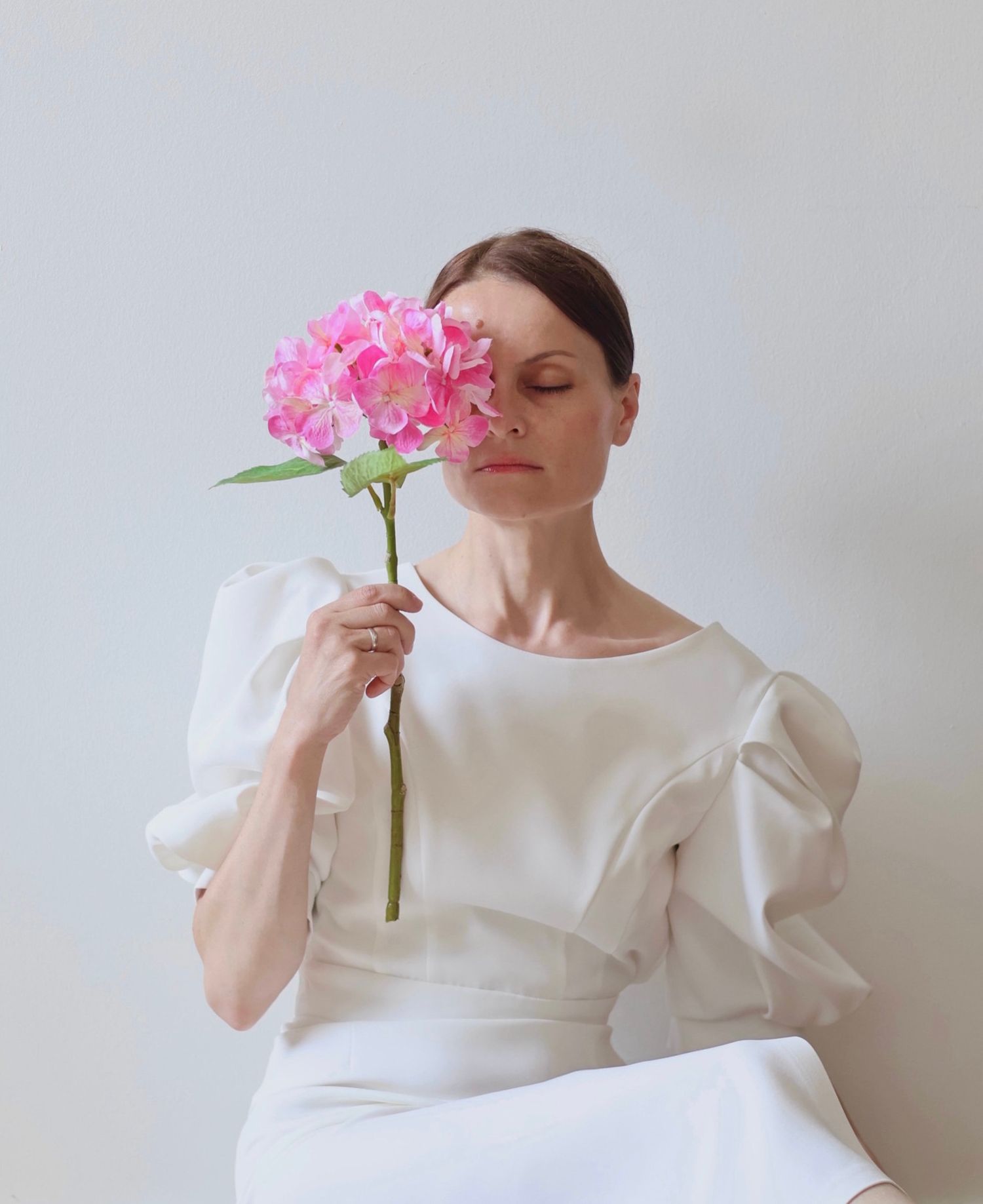 Woman in white puff-sleeve dress holds a pink hydrangea bloom in front of her face.
