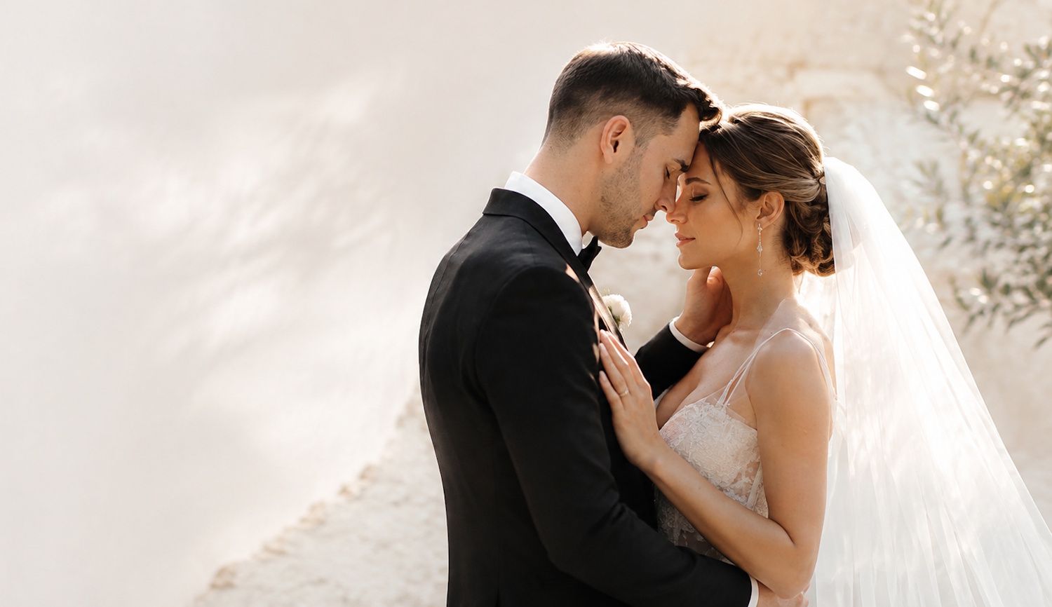 Bride and groom in wedding attire share an intimate forehead-to-forehead moment outdoors.