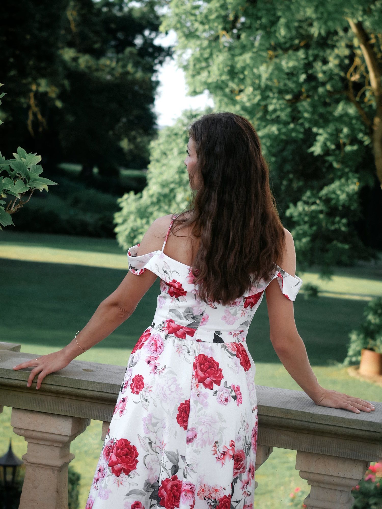 Woman in white floral dress viewed from behind on stone balcony in garden.