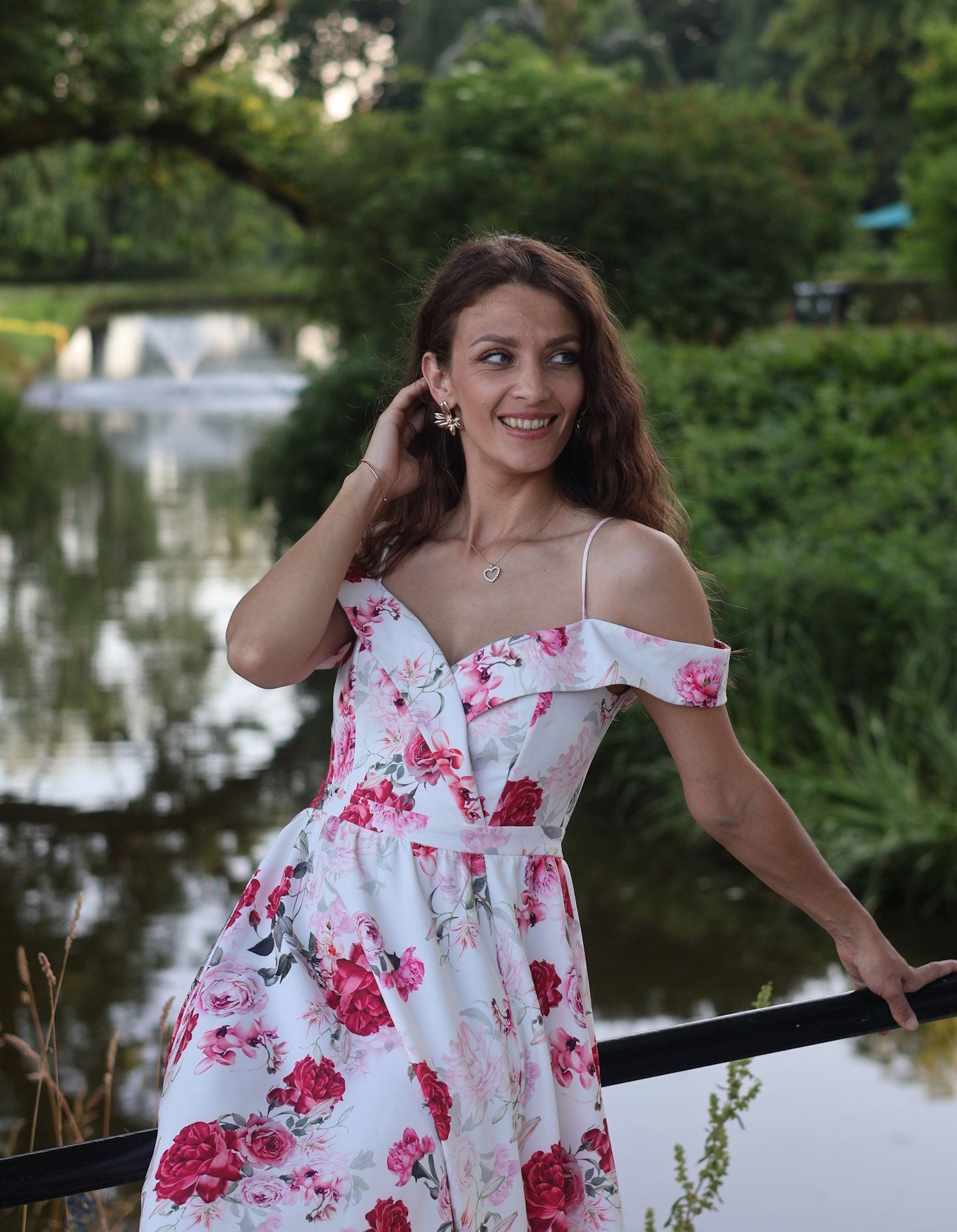 Smiling woman in floral off-shoulder dress posing by garden pond.