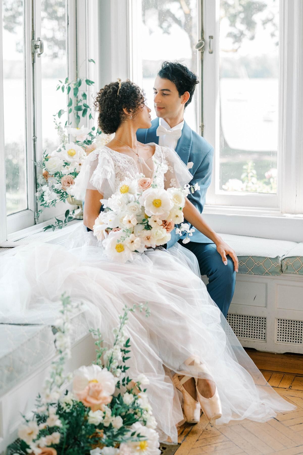Bride and groom share a romantic moment by a sunlit window, surrounded by white floral arrangements.