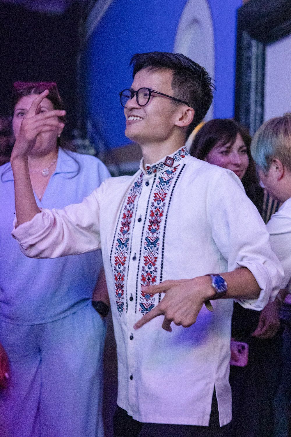 Young man in embroidered white shirt and glasses smiling and dancing at a social event.