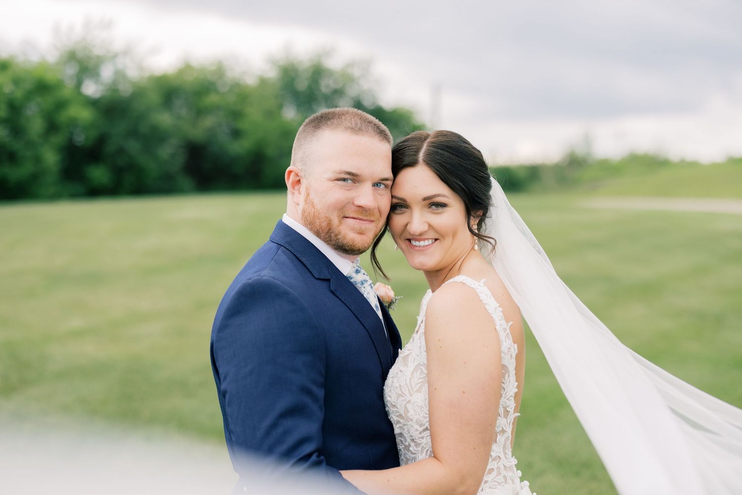 A wedding couple embraces outdoors in a romantic countryside setting with soft natural lighting and rolling hills in the background.
