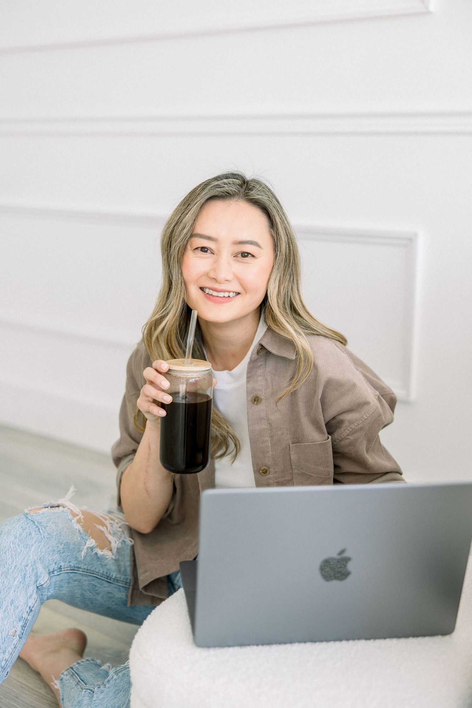 Someone in a brown jacket works on a MacBook laptop while enjoying an iced coffee drink at home.