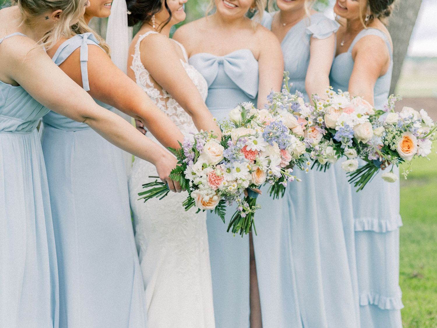 Bridesmaids in light blue dresses hold matching blush and white floral bouquets while standing in a row outdoors.