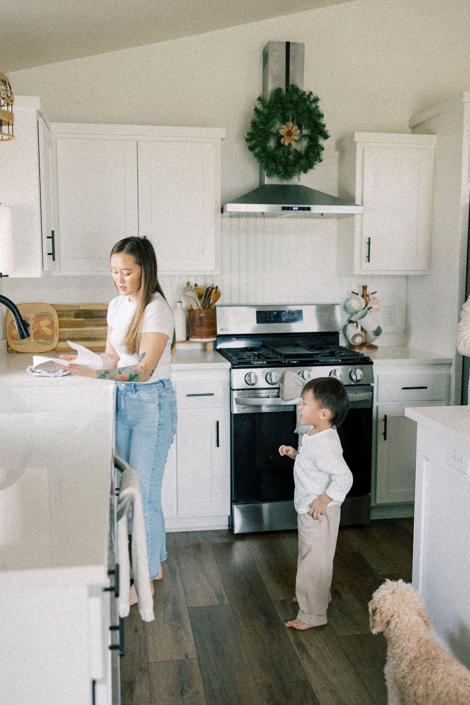Modern white kitchen with holiday wreath above range, featuring dark wood floors and a small fluffy dog.