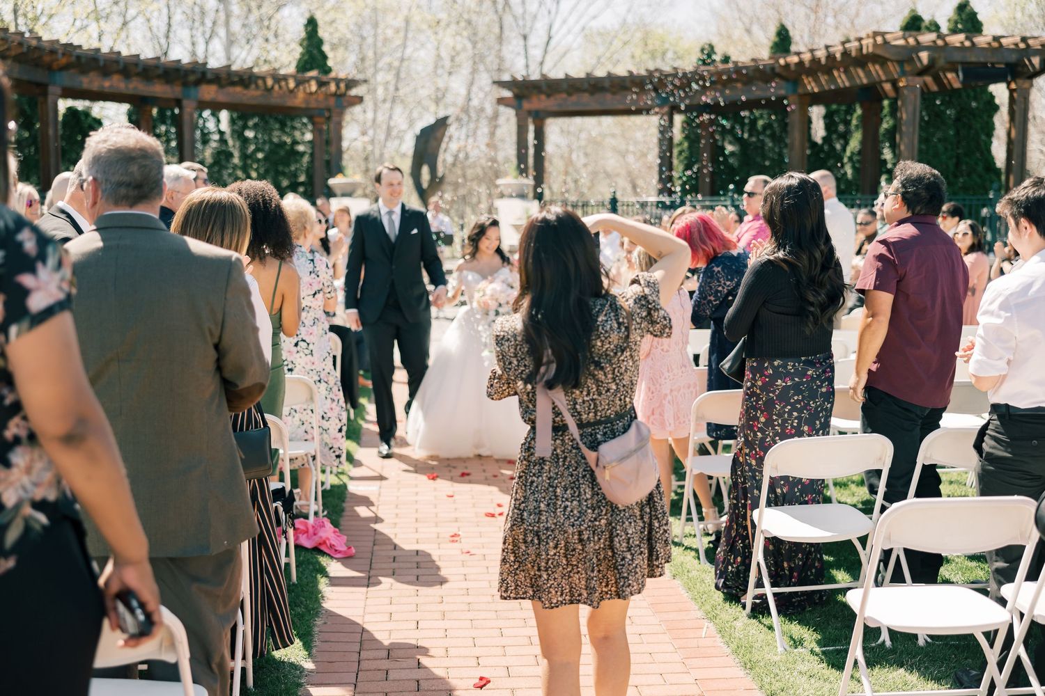 Guests gather outdoors at a garden wedding ceremony under a wooden pergola with white folding chairs and natural decor.