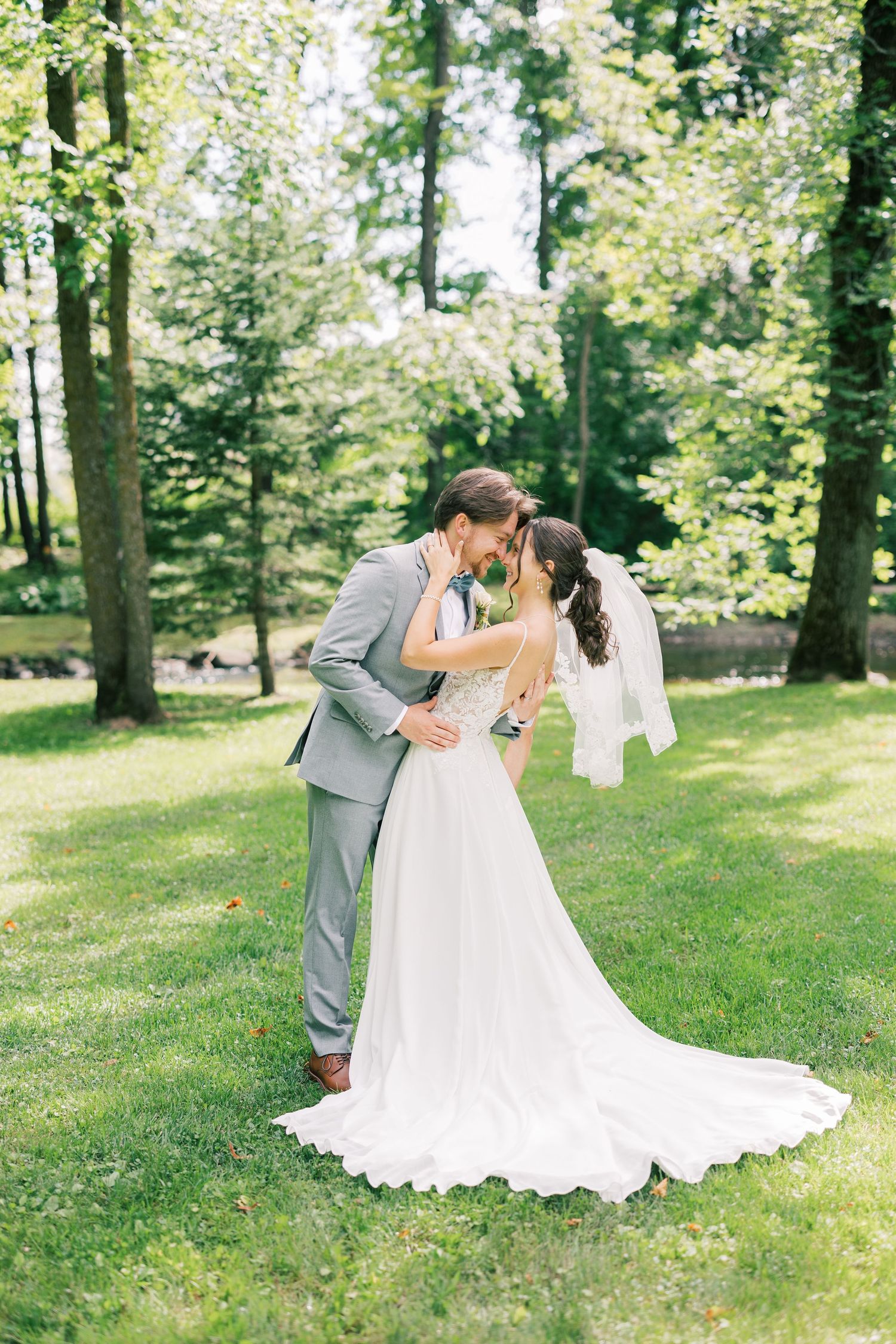 Romantic wedding couple shares intimate dips and kisses in a sunlit forest setting with white lace gown and gray suit.