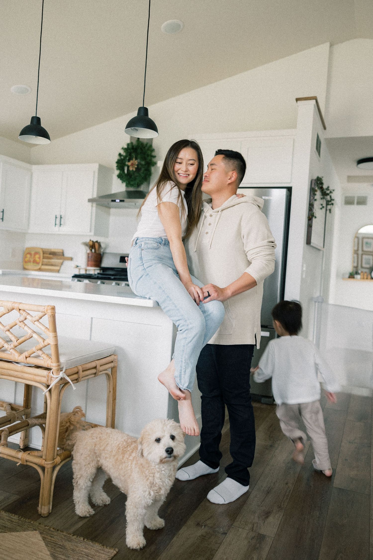 A couple sharing a tender moment in a bright modern kitchen with a white dog and child in the background.