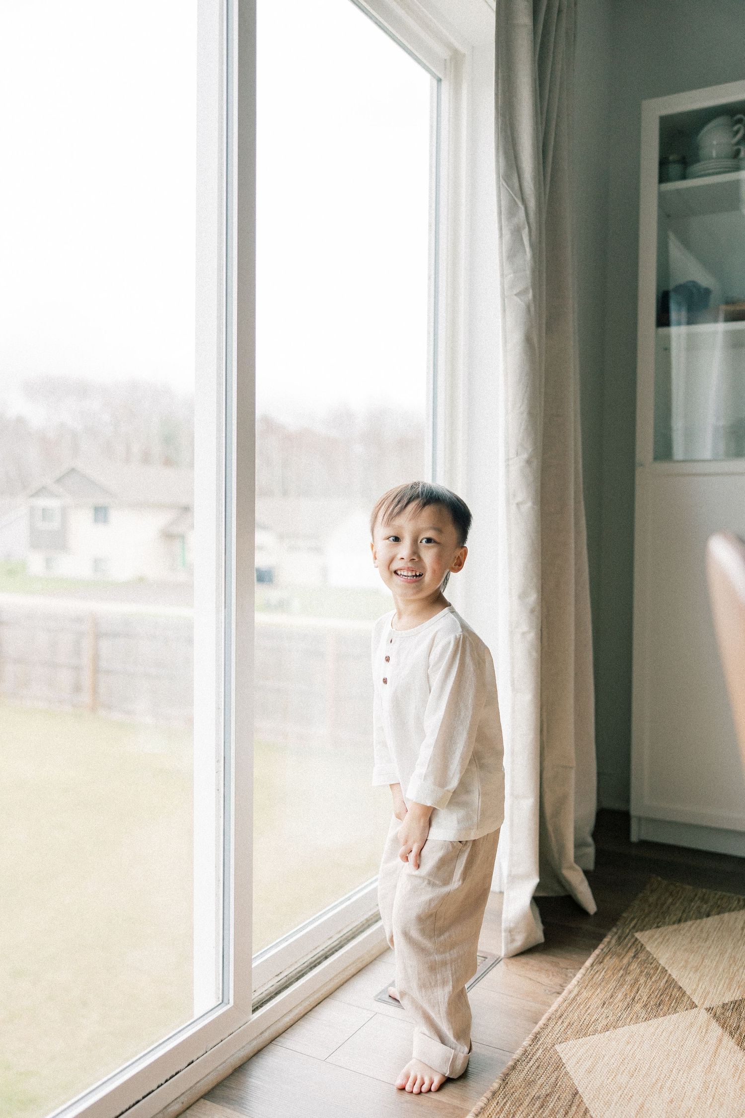 Little child in white clothing smiling while standing by a large window with natural light.