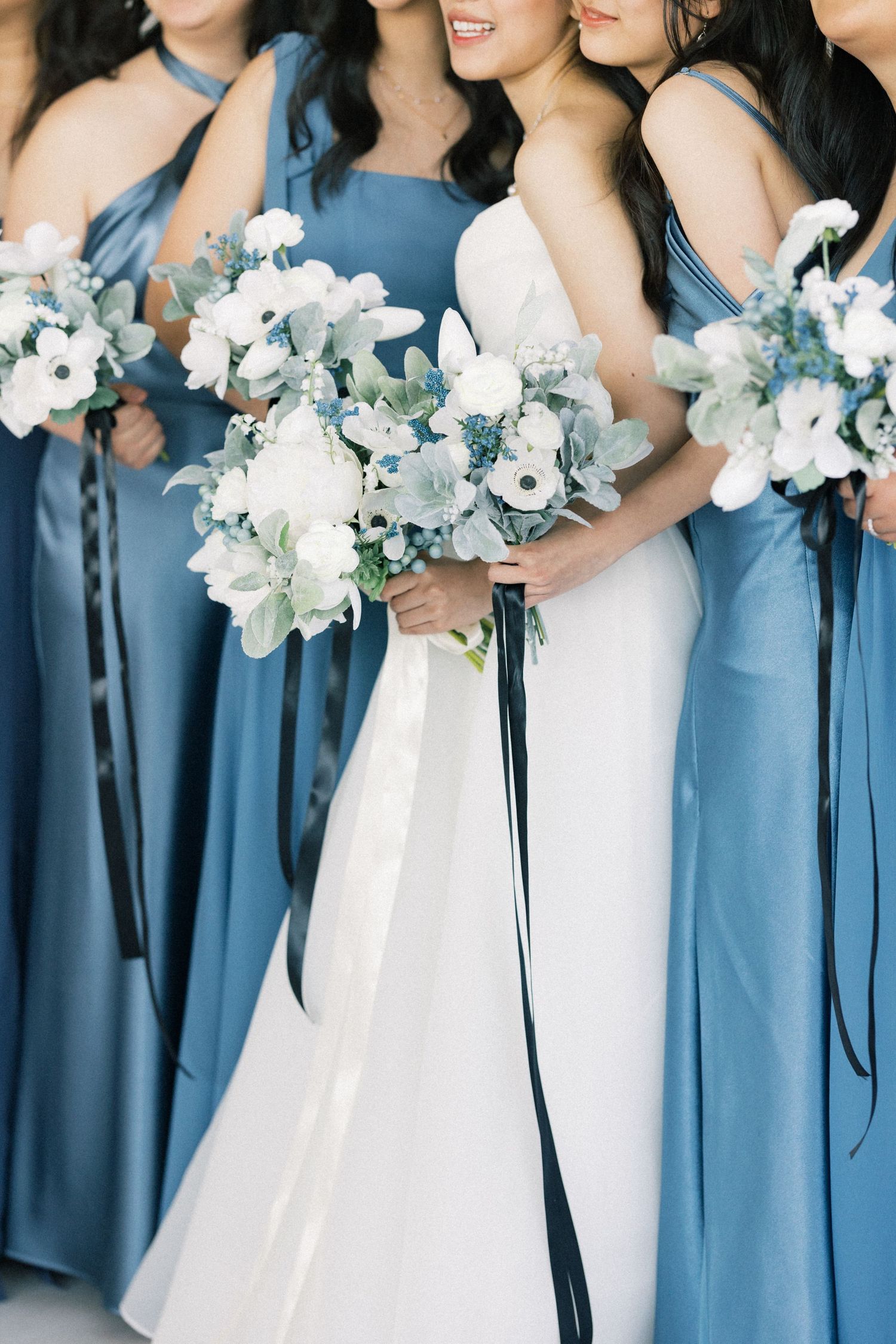 Bridesmaids in dusty blue dresses stand together holding white floral bouquets at an elegant wedding ceremony.