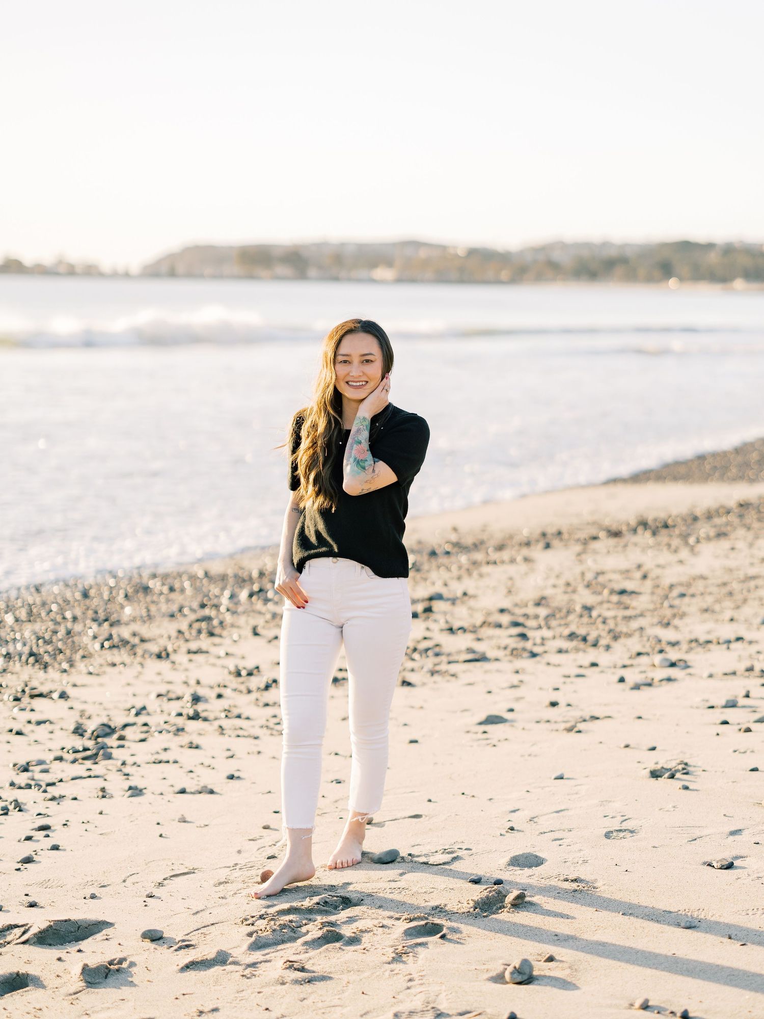 Person in black sweater and white pants standing on a rocky beach shore on a bright sunny day.