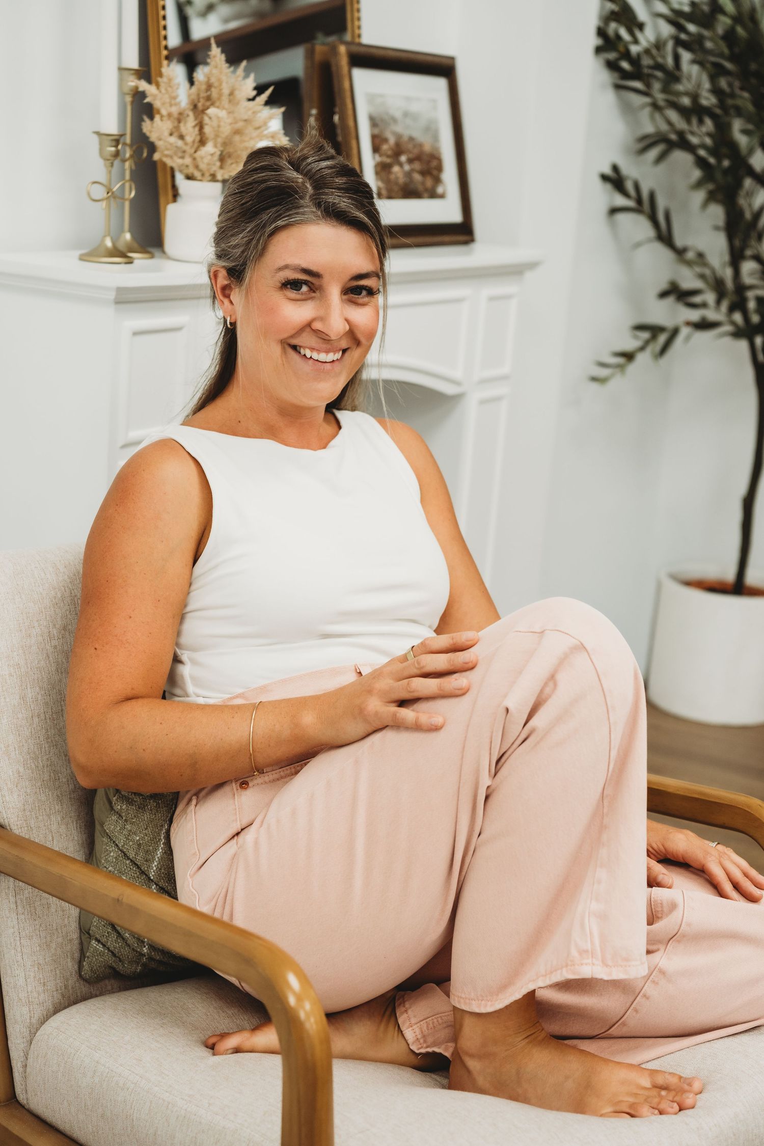 Smiling woman in white tank top and pink pants sitting cross-legged in a chair in a bright living room.