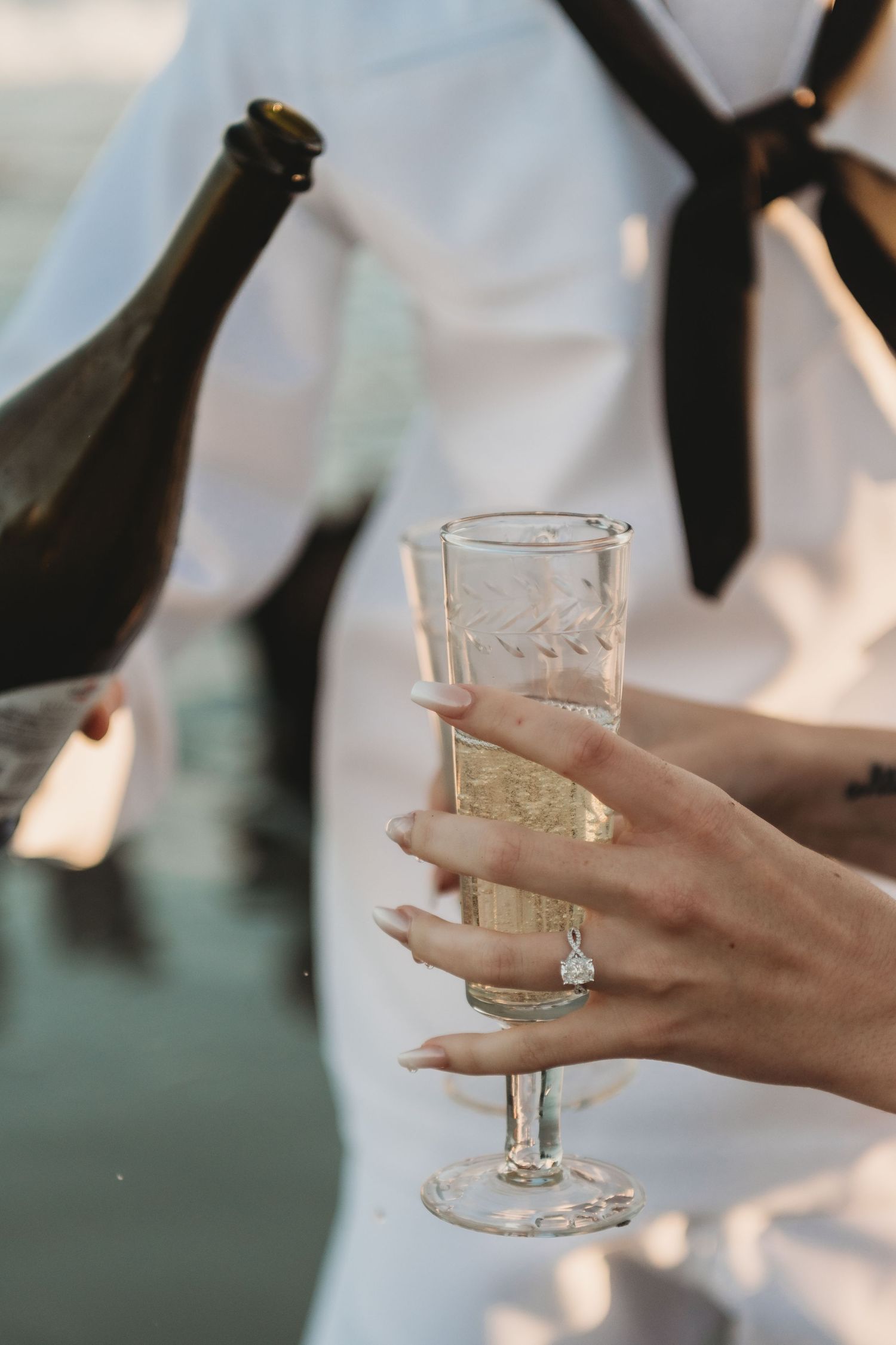 Close-up of womans hand with engagement ring holding champagne flute being filled at waterside.