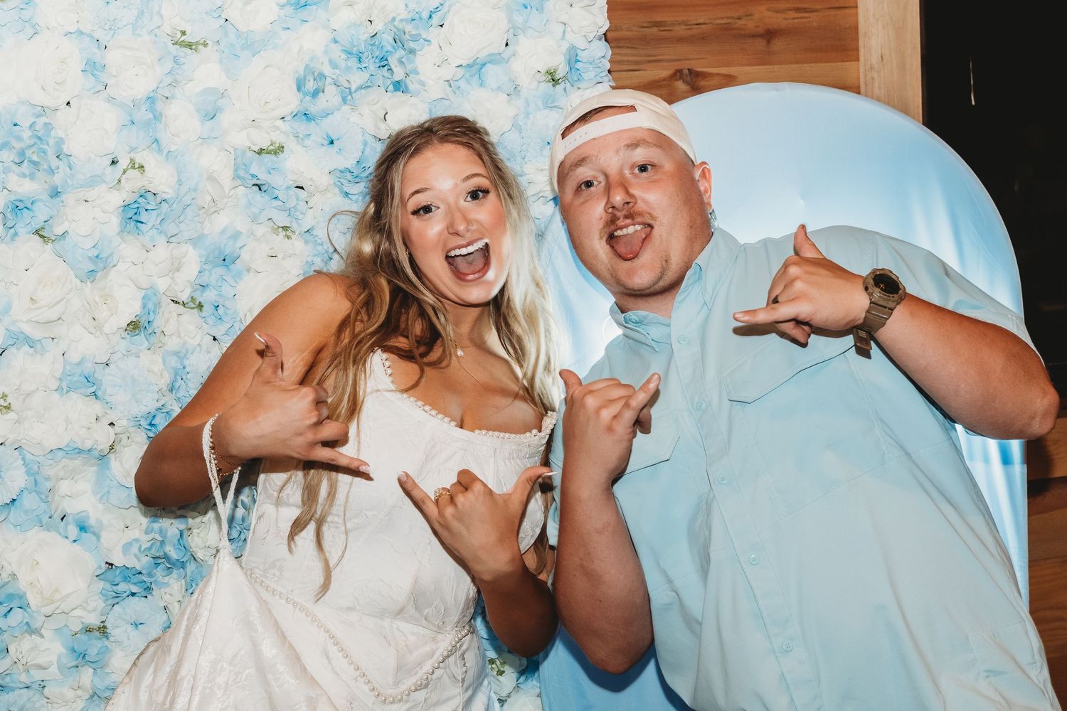 Bride and guest making shaka signs in front of blue and white floral wall at wedding reception.