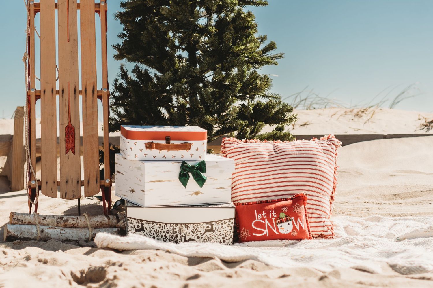 Christmas gifts and pillows arranged near a sled and tree on snowy beach.