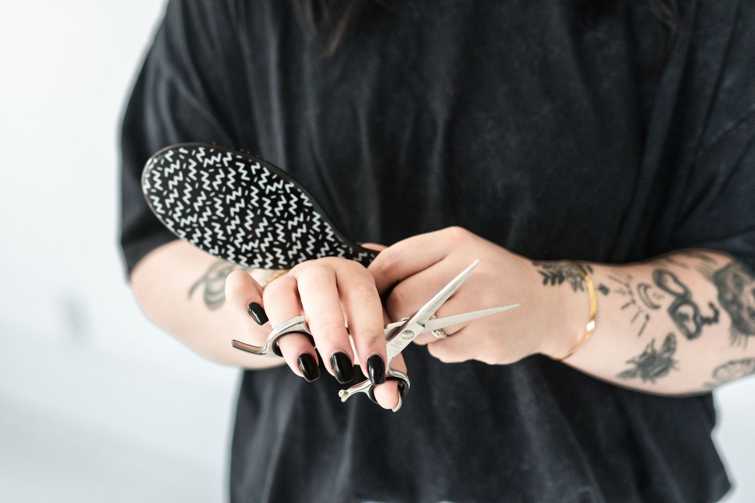 Tattooed hands holding hair brush and scissors with black nail polish in studio.